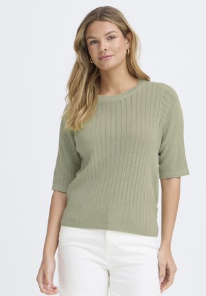 Naine helepruunide juustega, kandes kuldseid rõngaskõrvarõngaid, helerohelist soonikutega poolpika varrukaga kampsunit ja valgeid pükse, seismas neutraalse tausta ees.