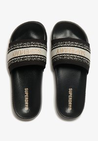 Chanclas negras con una correa texturizada que presenta un patrón decorativo y el logo "SUPERDRY" en dorado. Plantilla suave y acolchada.