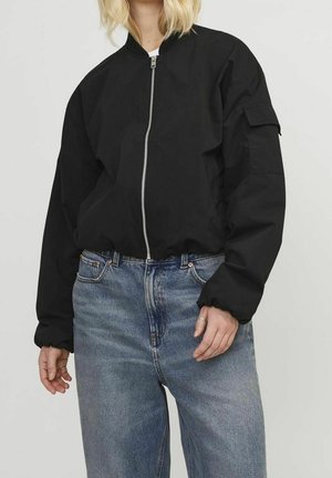 Person iført en sort bomberjakke med lynlås og en lomme på ærmet samt løstsiddende blå jeans foran en ensfarvet baggrund.