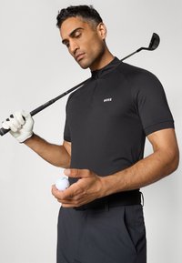 Svart golfpolo med korte ermer, glidelås i kvart lengde og strukturert finish; holder en hvit golfball og en golfklubbe, med en golfhanske på den ene hånden.