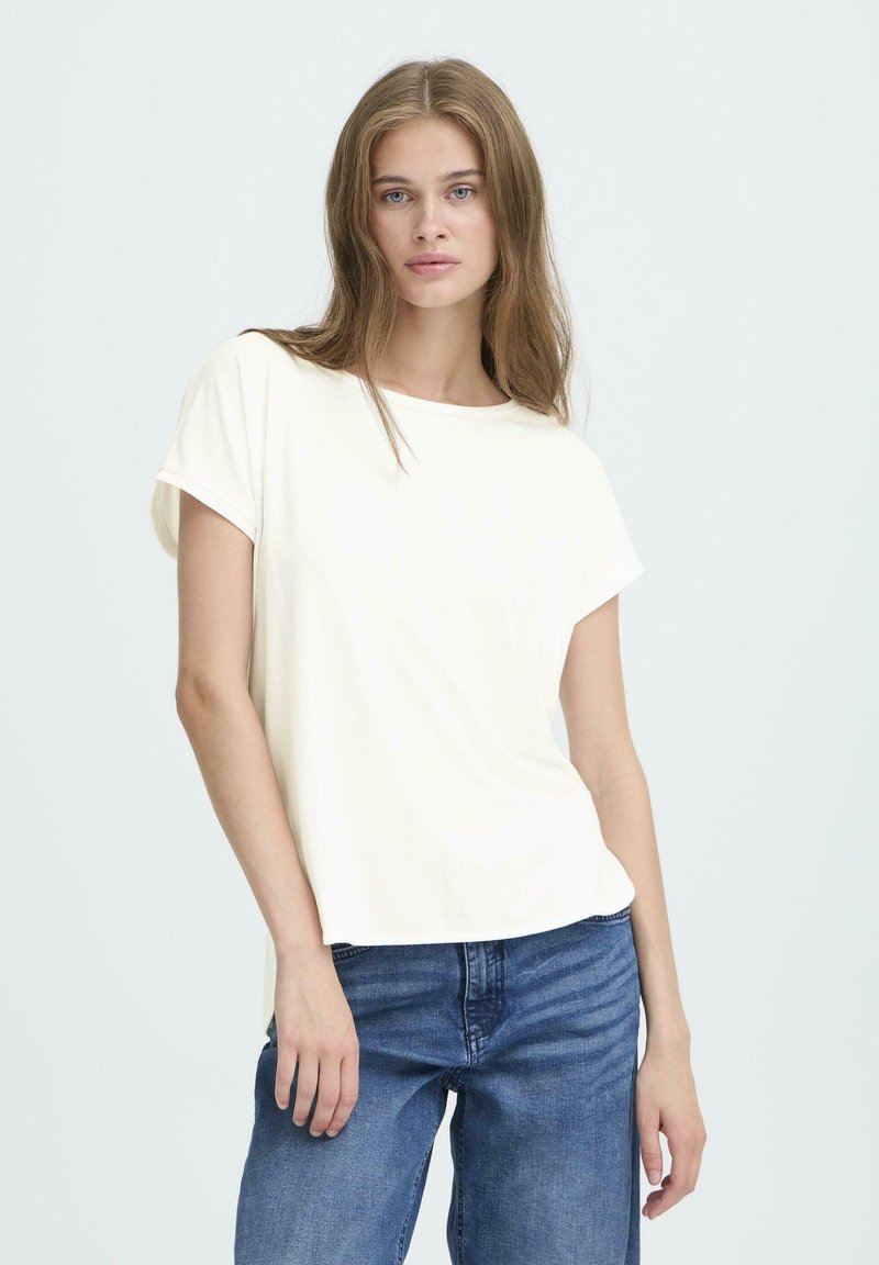 Ung kvinde med langt lysebrunt hår iført en løs hvid t-shirt med korte ærmer og blå jeans, der står foran en ensfarvet lys baggrund.
