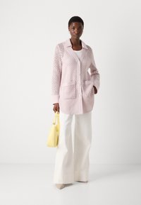  Lichtroze gebreide cardigan met een geperforeerde structuur, voorzien van een kraag, een knopenfront en twee voorzakken. Gecombineerd met witte broeken en een gele tas.
