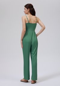 Combinaison verte avec un corsage ajusté et des jambes amples et longues. Fabriquée à partir d'un tissu lisse, elle possède des bretelles fines et ajustables ainsi qu'une taille cintrée.
