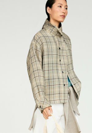 Personne portant une chemise à carreaux ample avec boutons et col montant, tenant un sac en tissu clair sur un fond uni.