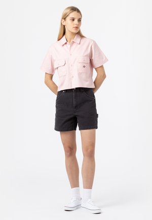 Afgebroken roze shirt met knoopsluiting en twee voorzakken, gecombineerd met zwarte denimshorts en witte sneakers.