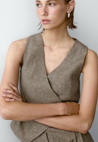 Brązowy lniany top bez rękawów o kroju kopertowym, z dekoltem w kształcie litery V i widocznymi detalami szwów. Gładka tekstura, minimalistyczny styl.