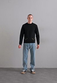 Bărbatul stă în fața unui zid simplu, purtând un pulover negru cu detalii din lanț pe buzunare, blugi bleu deschis și adidași negri cu șireturi albe.