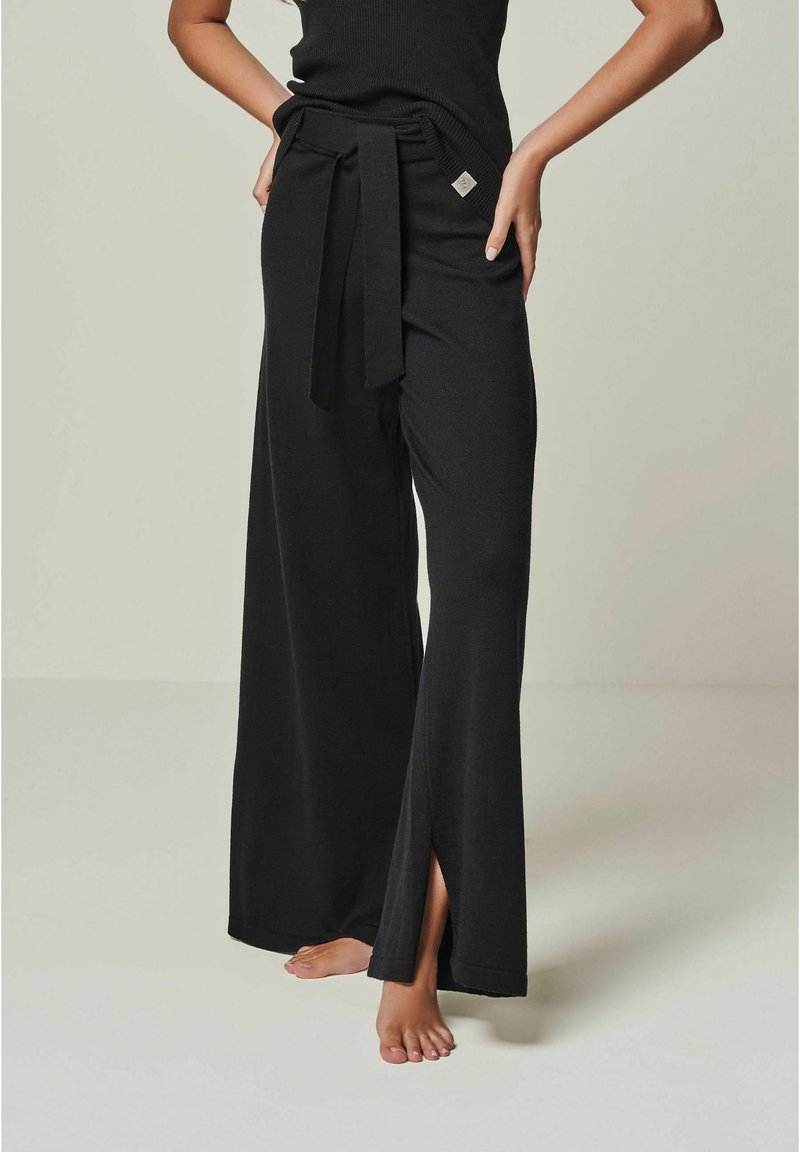 Schwarze weit geschnittene Hose mit einem taillierten Gurt, seitlichen Schlitzen und einer weichen Stoffstruktur. Mit einem kleinen Logodetail am Bund.