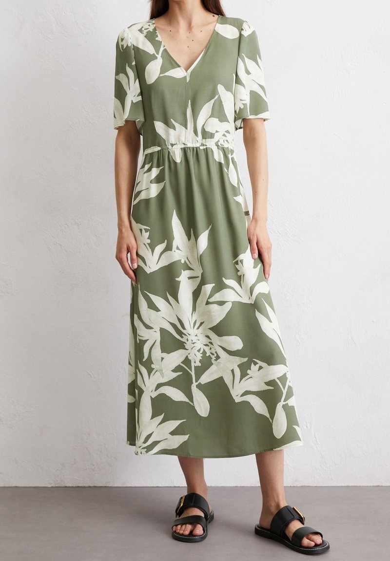 Blomstermønstret, knælang grøn kjole med korte ærmer, V-hals og elastisk talje. Kombineret med sorte flade sandaler med en gylden spænde.