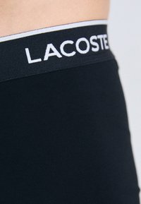 Zwarte rekbare stof tailleband met het "LACOSTE" logo in het wit, gladde textuur, aangesloten ontwerp, gedeeltelijk zicht op de shorts.