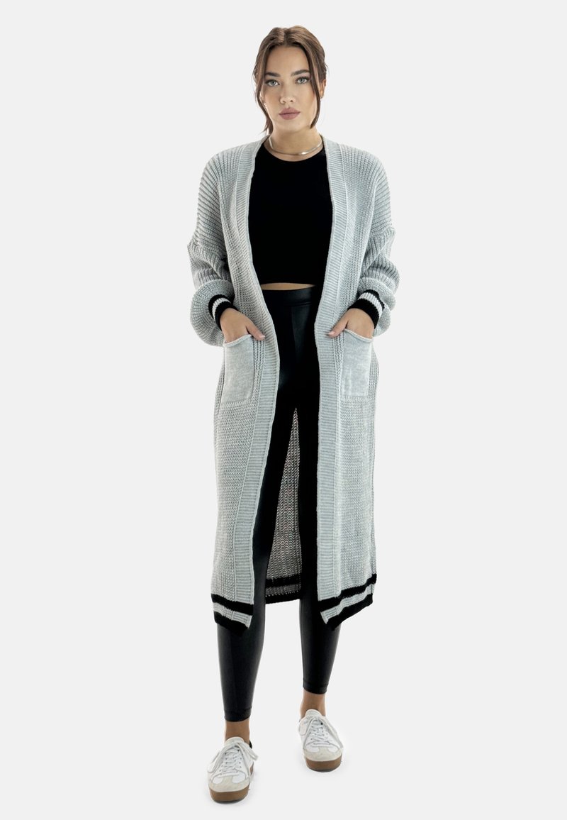Langer, hellgrauer Cardigan mit ribbedem Textur, schwarzen Akzenten und zwei vorderen Taschen, kombiniert mit schwarzen Leggings und weißen Turnschuhen.