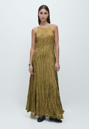 Kvinde med glat brunt hår iført en ærmeløs, krøllet, olivengrøn maxikjole og sorte sko, stående mod en ensfarvet lys baggrund.