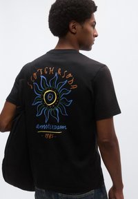 Μαύρο βαμβακερό T-shirt με πολύχρωμο γραφικό ήλιο στο πίσω μέρος, με εκτυπωμένα τα λόγια "Scotch & Soda," "Amsterdam" και "1985" σε αντίθετα χρώματα.