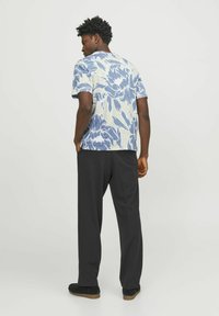 Man som står med ryggen vänd, iklädd en blå och vit blommig kortärmad skjorta, svarta byxor och svarta skor mot en enfärgad bakgrund.