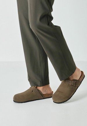 Brown suede clogs with a cross-strap design, adjustable buckle, and textured black rubber sole, paired with olive-green pants.
