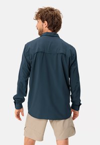 Camicia a maniche lunghe in tessuto blu scuro, con yoke posteriore e polsini con bottoni, abbinata a pantaloni corti beige chiaro.