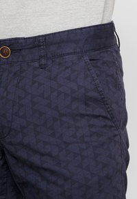 Pantaloni blu navy con un sottile motivo a triangoli scuri, dotati di tasca laterale, passante per cintura e bottone marrone in vita.