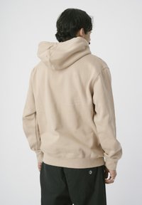 Beige Kapuzenpullover aus weichem Stoff mit Kängurutasche und Kordelzugkapuze. Verfügt über gerippte Ärmelbündchen und Saum, getragen mit dunklen Hosen.