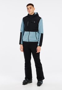 Zwarte en blauwe fleecejack met capuchon, rits aan de voorkant en details van zakken, gecombineerd met zwarte broek en skibotten. Glad textuur.