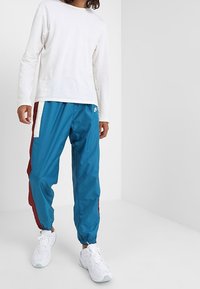 Person klädd i en vit långärmad skjorta, blå och vinröda träningsbyxor med vit rand, och vita sneakers som står mot en enhetlig bakgrund.