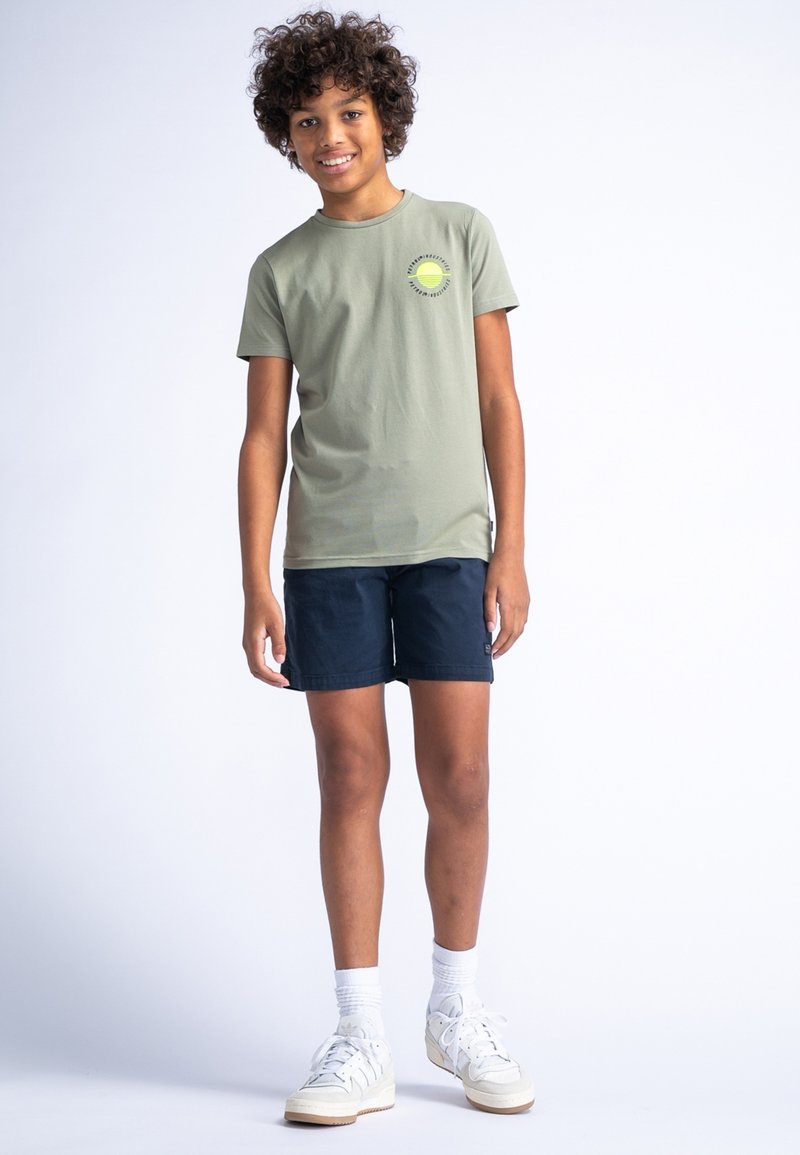 Groen katoenen t-shirt met korte mouwen en een rond logo, gecombineerd met marineblauwe shorts en witte sneakers. Licht, casual materiaal.