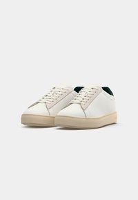 White sneakers with a smooth leather upper, beige accents, round toe, flat laces, and a textured rubber sole, featuring a minimalist design.
