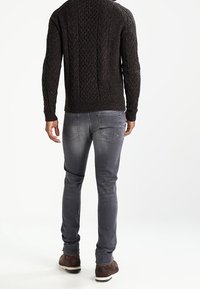 Pull en maille sombre avec un motif texturé, associé à un jean gris slim et des chaussures marron. Les détails visibles incluent des coutures de poche.