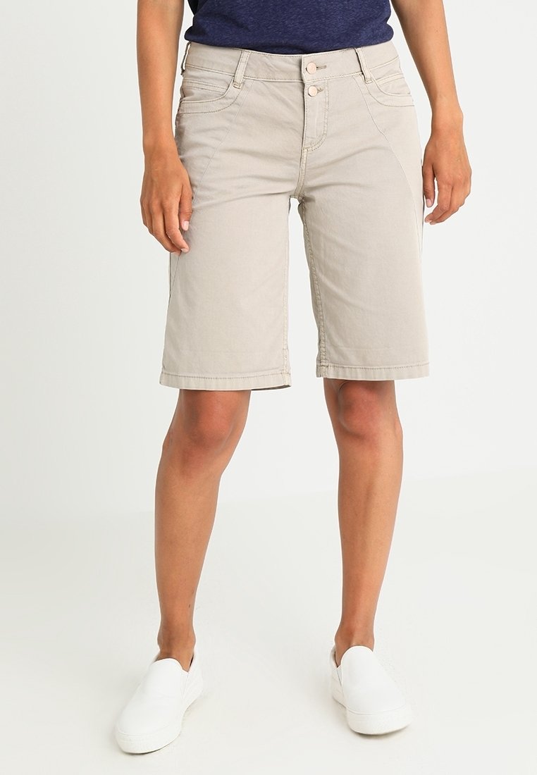 Shorts beige longueur genou avec poches à l'avant, portés avec un haut foncé et des chaussures blanches sans lacets, vus de la taille aux pieds.
