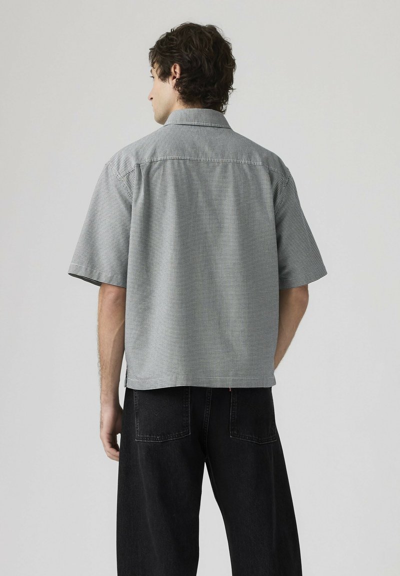 Uomo di spalle che indossa una camicia grigia a maniche corte con motivo e pantaloni neri larghi su uno sfondo chiaro semplice.