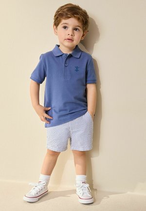 Ung dreng står mod en beige væg iført blå polo, stribede shorts, hvide sokker og hvide sneakers med hænderne i lommerne.