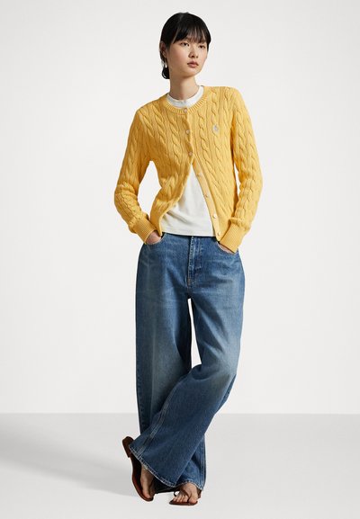 Gelber Strickcardigan mit rundem Hals und Knopfverschluss. Getragen über einem weißen T-Shirt. Kombiniert mit lockeren blauen Jeans und braunen Sandalen.