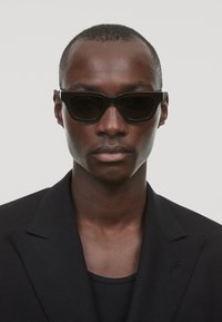 Mustad mustad prillid nurkadega raamiga, tumedate läätsede ja siledat viimistlust. Kandnud mudel musta bleiseri all tumeda särgi peal.