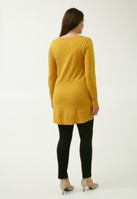 Maglione giallo senape a maniche lunghe con scollo rotondo sul retro, orlo a strati, abbinato a leggings neri e tacchi beige scintillanti.