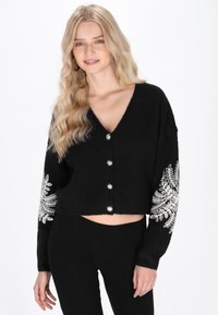 Čierny skrátený cardigan s V-výstrihom, strieborným predným zapínaním a podrobnou bielou výšivkou na rukávoch. Mäkká textura, voľný strih.