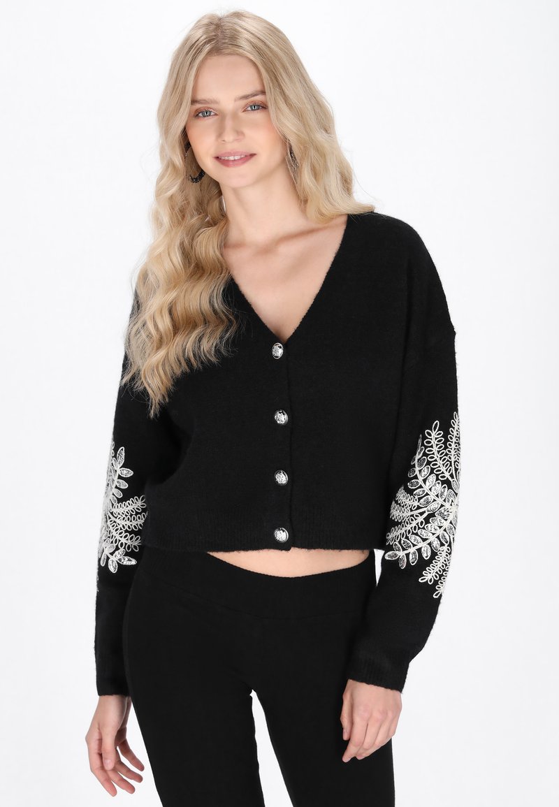 Čierny skrátený cardigan s V-výstrihom, strieborným predným zapínaním a podrobnou bielou výšivkou na rukávoch. Mäkká textura, voľný strih.
