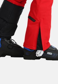Rote Skihose mit schwarzem unteren Abschnitt, seitlichen Reißverschlüssen; schwarze Skischuhe mit blauen Akzenten und weißen Schnallen.