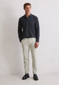 Mężczyzna w granatowej koszuli polo z długim rękawem, be�żowych spodniach, czarnym pasku i czarnych mokasynach stoi przy gładkiej białej ścianie.