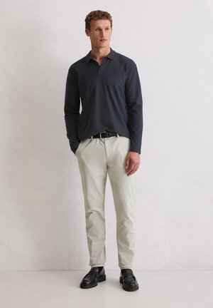 Homme portant un polo à manches longues bleu marine, un pantalon beige, une ceinture noire et des mocassins noirs, debout devant un mur blanc uni.