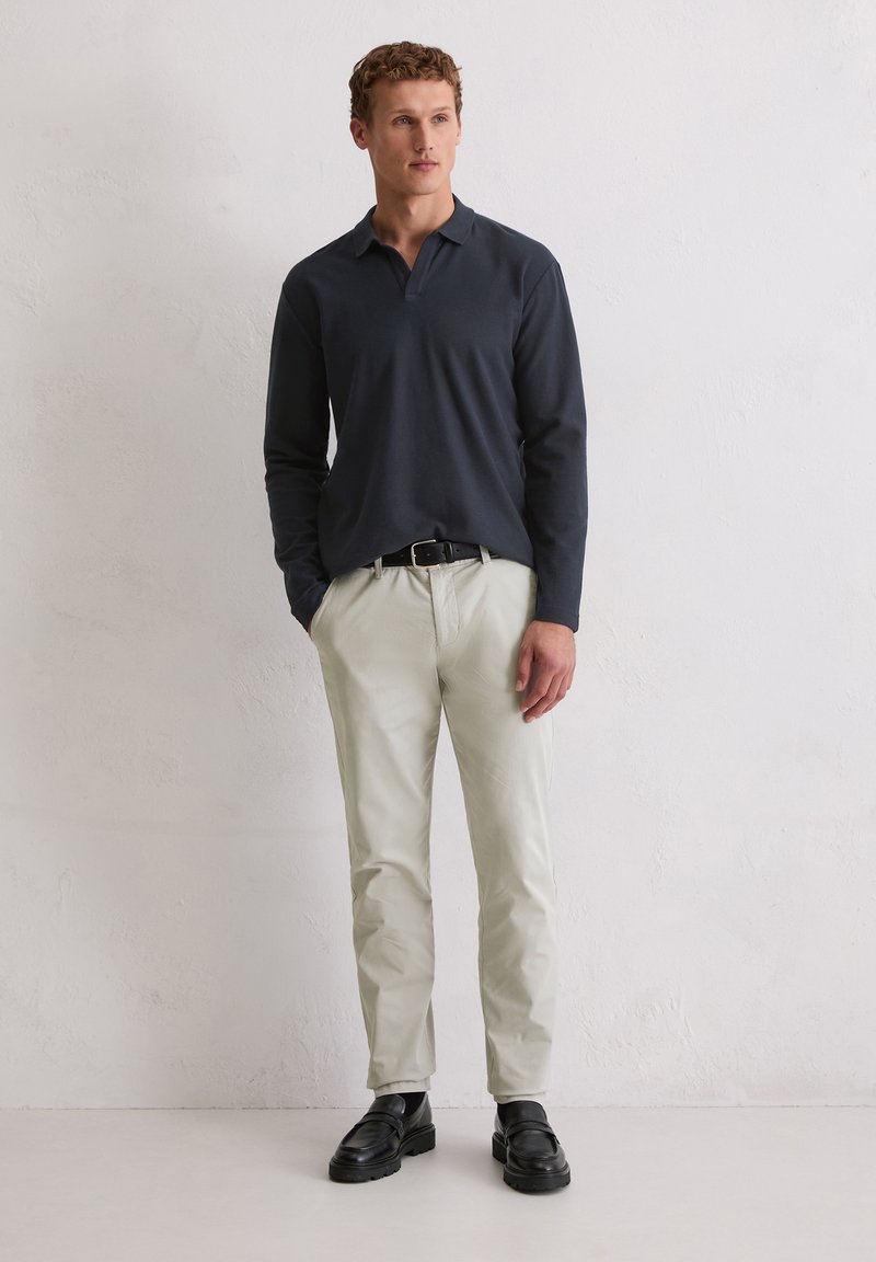 Mężczyzna w granatowej koszuli polo z długim rękawem, beżowych spodniach, czarnym pasku i czarnych mokasynach stoi przy gładkiej białej ścianie.