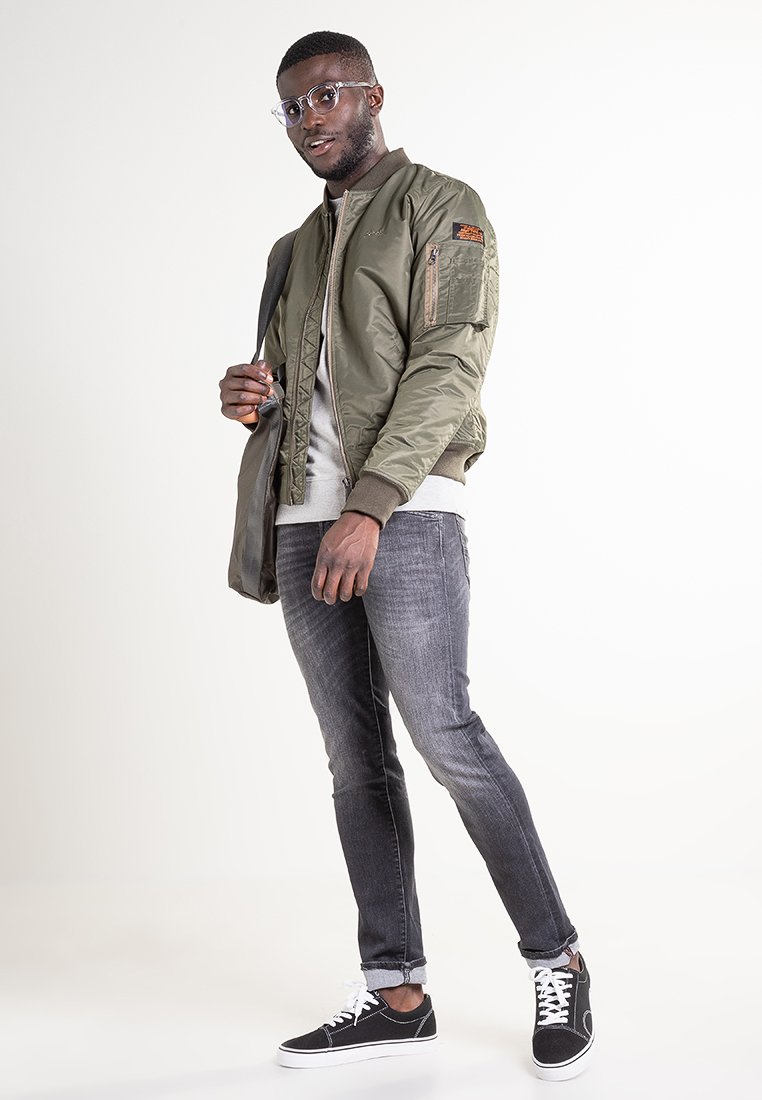 Veste bomber vert olive avec des poignets côtelés, sweat gris, jean slim gris, et baskets noires. Lunettes noires et sac à bandoulière inclus.