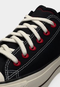 Μαύρο αθλητικό παπούτσι με λευκά κορδόνια που περνούν μέσα από κόκκινα οπές και λεπτομερή λευκή ραφή πάνω στο υφασμάτινο επάνω μέρος και μπεζ καουτσούκ σόλα.