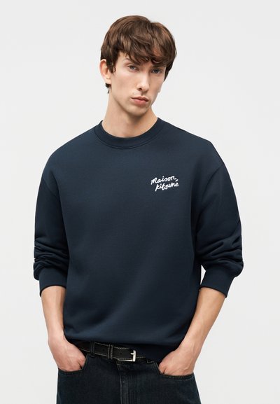 Νέος με καστανά μαλλιά που φοράει μπλε φούτερ με το κείμενο "Maison Kitsuné", χέρια στις τσέπες, σε απλό ανοιχτό φόντο.