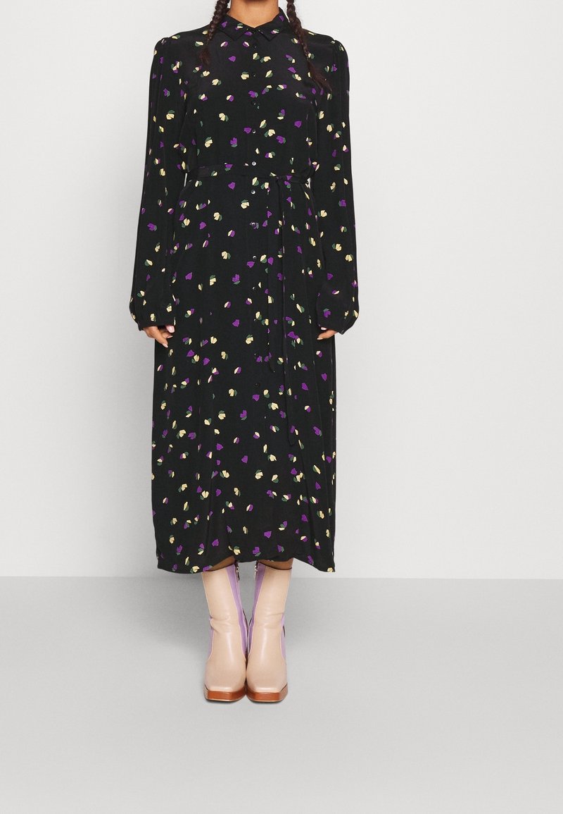 Femme portant une robe midi noire à manches longues avec un imprimé floral violet et jaune, associée à des bottines rose clair.