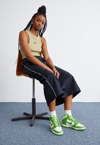 Green and white high-top Nike sneakers, ribbed olive tank top, black wide-leg pants with white piping, seated on a swivel chair.