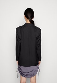 Sort blazer med strukturerede skuldre, tekstureret stof og lige snit, parret med en draperet lilla kjole med bånddetaljer.
