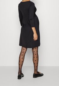 Svart långärmad klänning med en figurnära bodice och utsvängd kjol. Bärs med genomskinliga svarta strumpbyxor med prickar och svarta platta skor med guldaccenter.