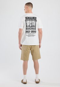 White t-shirt featuring a black graphic print of a café storefront with the text "DAILY GRIND," paired with beige shorts and slippers.
