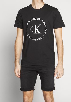 T-shirt en coton noir avec un col rond, présentant un logo circulaire blanc avec les lettres "CK" et le texte "Calvin Klein Jeans". Design simple, manches courtes.