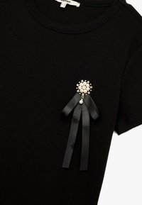 Camiseta de algodón negra con un broche decorativo hecho de perlas y acentos metálicos, con cintas de satén negro unidas.