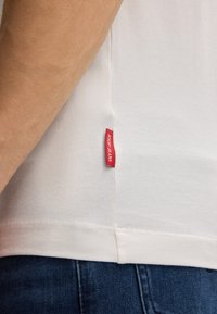 Nahaufnahme einer Person von der Seite, die ein weißes Hemd mit einem roten "Joop! Jeans"-Etikett und blaue Jeans trägt.